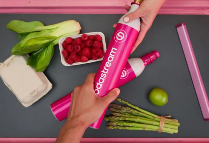 Hands swapping pink Sodastream CO2 cylinders over a counter of fresh groceries